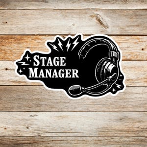 May include: A black and white sticker featuring the text STAGE MANAGER in a bold serif font, accompanied by an illustration of a headset with a microphone, surrounded by stylized lightning bolt and star accents on a die-cut white border.