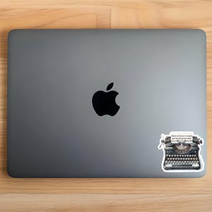 Op de afbeelding: Een donkergrijze laptop met een zwart Apple-logo in het midden. Een sticker van een zwarte vintage typemachine is in de rechterbenedenhoek geplaatst, met een papierinzetstuk waarop staat, None of your e-mails are finding me well.