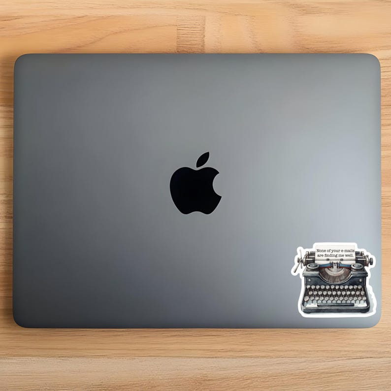 Op de afbeelding: Een donkergrijze laptop met een zwart Apple-logo in het midden. Een sticker van een zwarte vintage typemachine is in de rechterbenedenhoek geplaatst, met een papierinzetstuk waarop staat, None of your e-mails are finding me well.