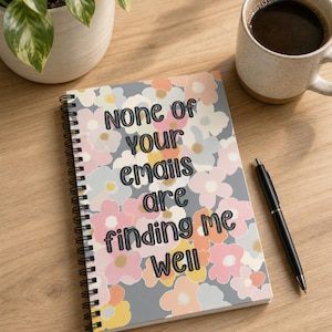 May include: A spiral-bound notebook featuring a floral pattern in shades of pink, yellow, light blue, and grey. The cover displays the text "NONE of your emails are finding me well" in a dark, rounded, bold font centered on the floral background.