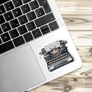 Op de afbeelding: Een sticker met een zwarte vintage typemachine met een vel papier waarop staat: "None of your e-mails are finding me well." De typemachine heeft gedetailleerde toetsen, een wagen en een metalen afwerking op een zilveren laptop.