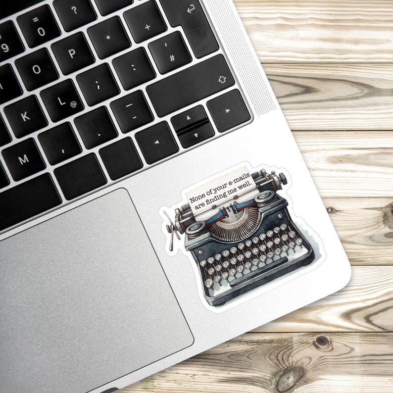 Op de afbeelding: Een sticker met een zwarte vintage typemachine met een vel papier waarop staat: "None of your e-mails are finding me well." De typemachine heeft gedetailleerde toetsen, een wagen en een metalen afwerking op een zilveren laptop.
