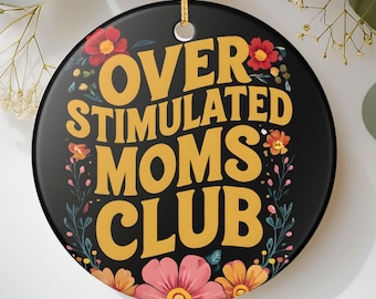 May include: A round black ceramic ornament featuring the yellow text OVER STIMULATED MOMS CLUB in a bold, wavy font. The design is decorated with colorful red, pink, and orange floral illustrations and green stems, hanging from a gold string.