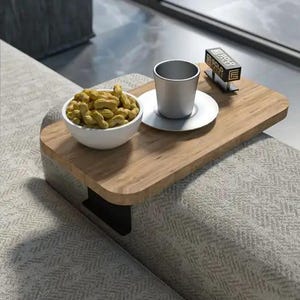 May include: A wooden sofa arm tray with a metal base holds a white bowl of peanuts, a silver cup and saucer, and a small box. The tray is attached to a grey patterned sofa arm.