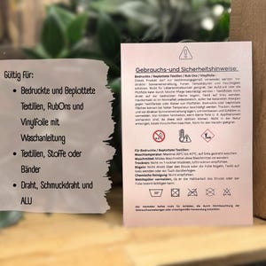 Può includere: Una scheda bianca con testo in tedesco che fornisce istruzioni per la cura e la sicurezza per tessuti stampati e vinile. La scheda include linee guida per il lavaggio, l'asciugatura e la stiratura, insieme ad avvertenze di sicurezza. La scheda ha anche un simbolo di temperatura.