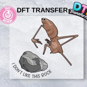 May include: A DTF Transfer design featuring a cartoon alien-like creature with long limbs and a rock. The text "I DON'T LIKE THIS ROCK" is below the rock. The image also includes a "Fast Delivery" logo.