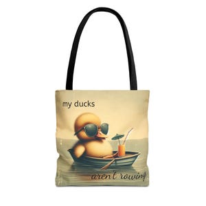 Puede incluir: Bolso tote con una ilustración de un pato amarillo con gafas de sol, relajándose en un bote con una bebida. El bolso tiene asas negras y el texto "my ducks aren't rowing".
