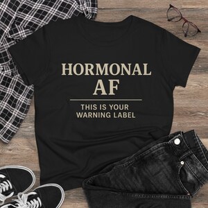 Könnte beinhalten: Schwarzes T-Shirt mit dem Text "HORMONAL AF" über einer Linie und "THIS IS YOUR WARNING LABEL" darunter. Das Shirt wird mit schwarzen Jeans, schwarz-weißen Turnschuhen und einer Brille präsentiert.