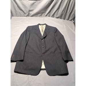 May include: A dark gray pinstripe blazer with a three-button closure. The blazer has a notched lapel and a light-colored lining. The sleeves have three buttons at the cuff. The blazer is laid flat on a light gray surface.