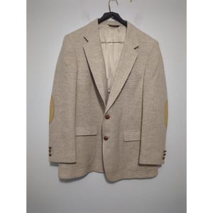 May include: A beige blazer with brown buttons and tan elbow patches hangs on a black hanger. The blazer has two front pockets and a notched lapel. The fabric has a textured appearance.