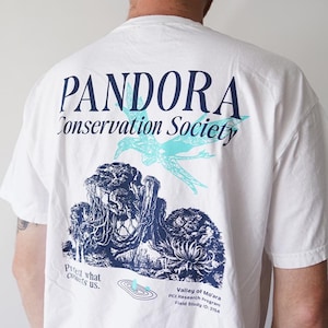 May include: White t-shirt with a graphic design featuring the text "PANDORA Conservation Society." The design includes a teal bird and a blue and white illustration of a landscape. Additional text includes "Protect what connects us" and "Valley of Mo'ara."
