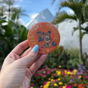 May include: A round orange pinback button featuring a cartoon trash can character with arms and legs, a signpost, and a circular object. The text reads FESTIVAL FOODIE and TURNING TRASHCANS INTO TABLES in a light yellow font against the orange background.