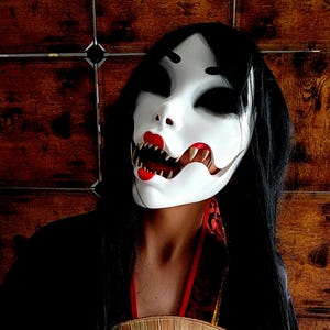 May include: A woman with long black hair wears a white mask with a red mouth and sharp teeth. She is holding a large, round, woven fan in front of her.
