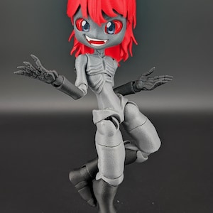 May include: A 3D-printed articulated figure of a stylized humanoid creature. The figure has a gray body, black limbs, and a red hairstyle. It features a smiling expression with red eyes and is posed with arms outstretched.