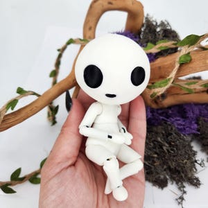 May include: A white articulated doll with a round head and large black eyes. The doll is sitting in a hand, with a wooden branch and greenery in the background. The doll's arms and legs are poseable.