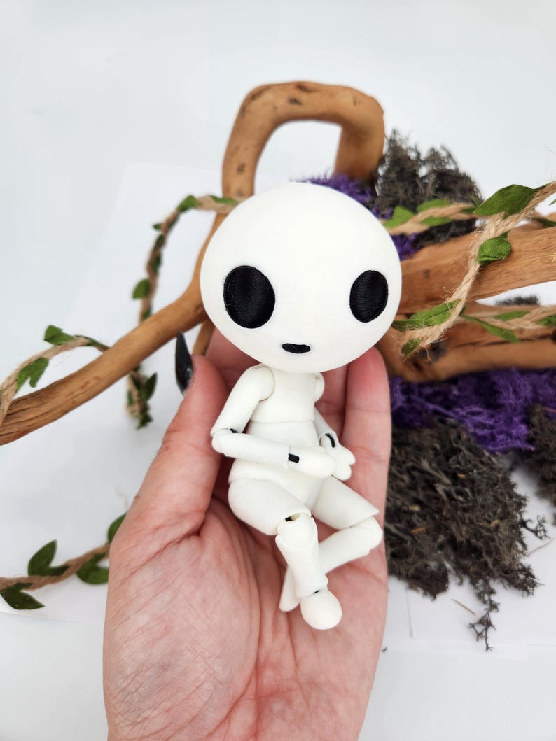 May include: A white articulated doll with a round head and large black eyes. The doll is sitting in a hand, with a wooden branch and greenery in the background. The doll's arms and legs are poseable.