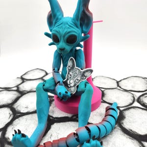 May include: A blue and red articulated cat figurine with a black and white cat print on its chest. The figurine is sitting on a pink stand and has a segmented tail.