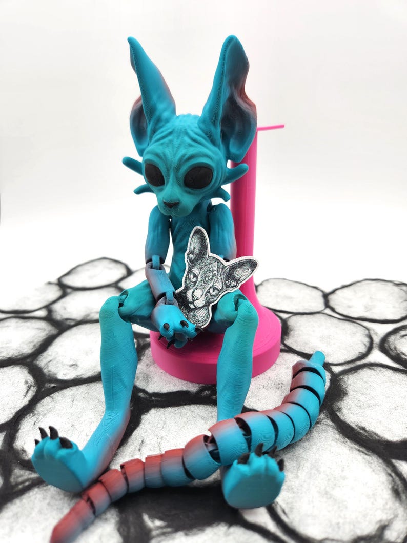 May include: A blue and red articulated cat figurine with a black and white cat print on its chest. The figurine is sitting on a pink stand and has a segmented tail.