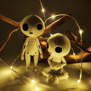 May include: Two white, articulated dolls with large black eyes and small mouths are illuminated by string lights. One doll stands, the other sits. A piece of driftwood is in the background, adding a natural element to the scene.