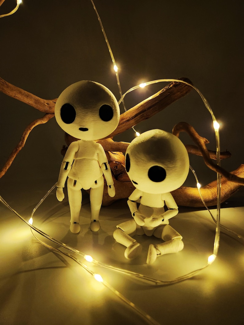 May include: Two white, articulated dolls with large black eyes and small mouths are illuminated by string lights. One doll stands, the other sits. A piece of driftwood is in the background, adding a natural element to the scene.