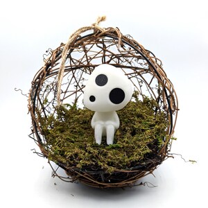 K&ouml;nnte beinhalten: Eine wei&szlig;e Figur mit drei schwarzen Punkten sitzt in einem kugelf&ouml;rmigen, gewebten Nest, das mit gr&uuml;nem Moos gef&uuml;llt ist. Das Nest besteht aus braunen Zweigen und wird von einem nat&uuml;rlichen Bindfaden gehalten. Die Figur hat eine pilzartige Form.