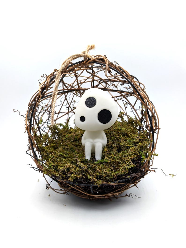 K&ouml;nnte beinhalten: Eine wei&szlig;e Figur mit drei schwarzen Punkten sitzt in einem kugelf&ouml;rmigen, gewebten Nest, das mit gr&uuml;nem Moos gef&uuml;llt ist. Das Nest besteht aus braunen Zweigen und wird von einem nat&uuml;rlichen Bindfaden gehalten. Die Figur hat eine pilzartige Form.