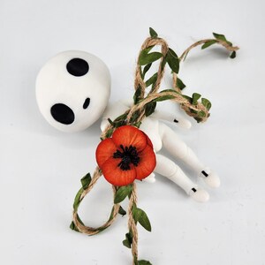 May include: A white doll with black eye markings and a red poppy flower. The doll is decorated with a twine vine with green leaves. The doll's arms and legs are white.