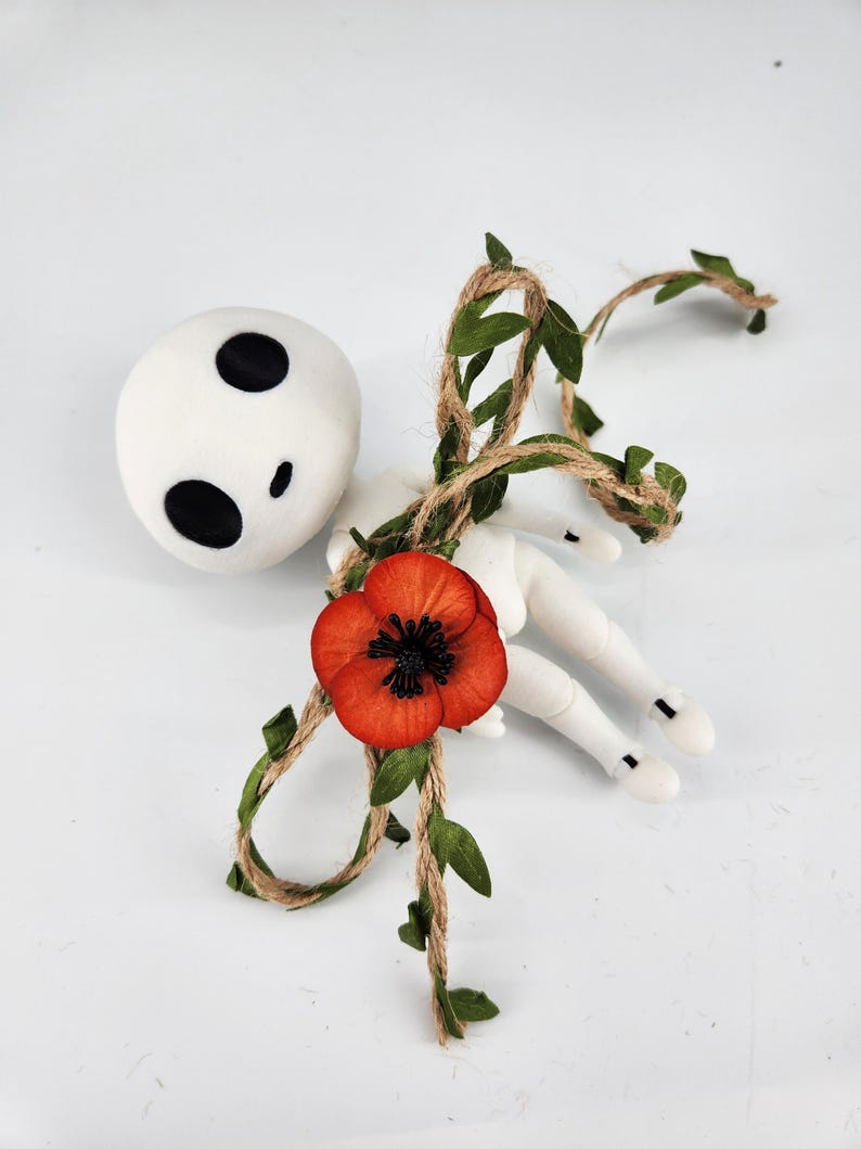 May include: A white doll with black eye markings and a red poppy flower. The doll is decorated with a twine vine with green leaves. The doll's arms and legs are white.