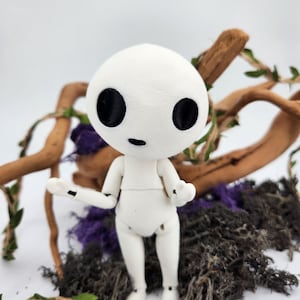 May include: A white articulated doll with a round head and large black eyes. The doll is posed with arms outstretched, standing on a bed of dark moss, purple accents, and wooden branches. The doll's design is reminiscent of a forest spirit.