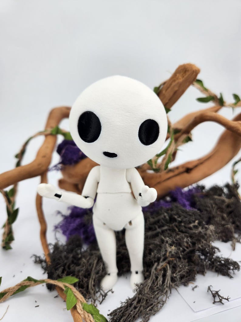 May include: A white articulated doll with a round head and large black eyes. The doll is posed with arms outstretched, standing on a bed of dark moss, purple accents, and wooden branches. The doll's design is reminiscent of a forest spirit.