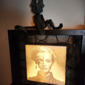May include: A black framed light box with a white and black image of a woman's face, half skull and half human, with a black skeletal figure with large ears sitting on top of the frame.