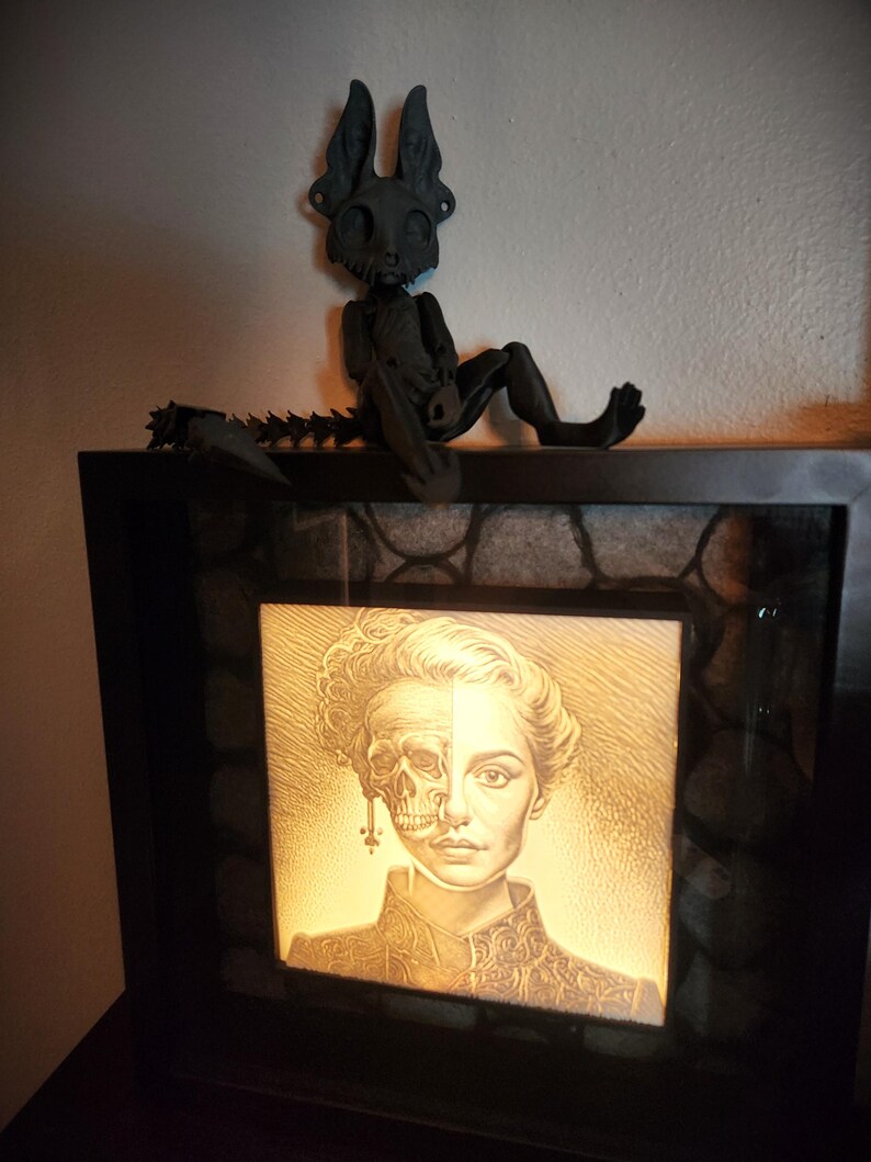 May include: A black framed light box with a white and black image of a woman's face, half skull and half human, with a black skeletal figure with large ears sitting on top of the frame.