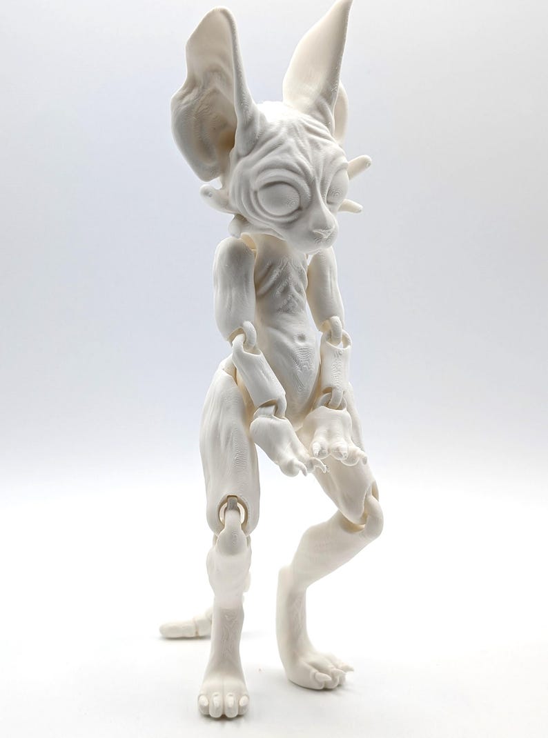 May include: White articulated doll with a cat-like face and large ears. The doll has a detailed, textured surface and is posed standing with its arms raised.
