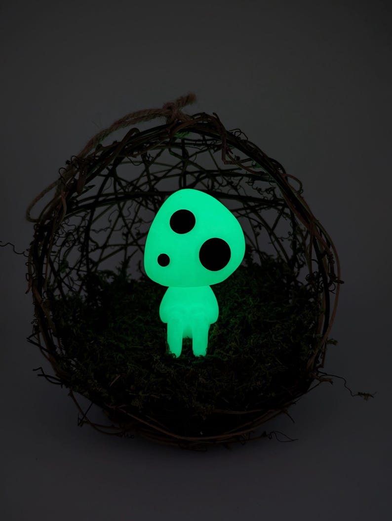 K&ouml;nnte beinhalten: Eine leuchtend gr&uuml;ne, pilzf&ouml;rmige Figur mit drei schwarzen, kreisf&ouml;rmigen Punkten. Die Figur befindet sich in einem gewebten, kreisf&ouml;rmigen Nest, das mit gr&uuml;nem Moos gef&uuml;llt ist. Der Hintergrund ist dunkel.