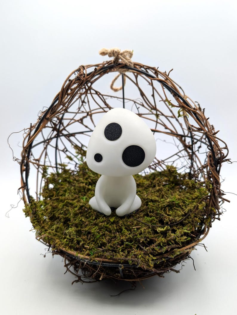 K&ouml;nnte beinhalten: Eine wei&szlig;e Figur mit drei schwarzen Punkten, die an einen Waldgeist erinnert, sitzt auf einem Bett aus gr&uuml;nem Moos in einer gewebten Zweigkugel. Die Kugel wird von einer nat&uuml;rlichen Schnurschlaufe gehalten, was ein skurriles Dekor ergibt.