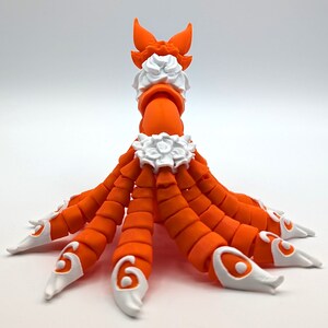 Peut inclure: Figurines orange et blanche d'une cr&eacute;ature &agrave; plusieurs bras et une queue. La cr&eacute;ature a un collier blanc et un motif floral blanc autour de la taille. La figurine est en plastique et a une finition lisse.
