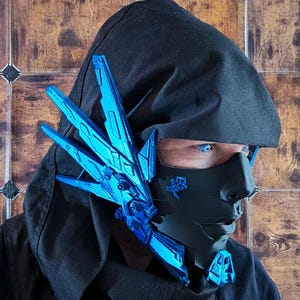 May include: A black face mask featuring a prominent, metallic blue mechanical wing-like attachment on the side. The mask includes a small blue symbol on the cheek area and a blue geometric accent on the chin, paired with a black hooded garment.