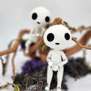 May include: Two white, articulated dolls with black eyes and small mouths, resembling characters from a fantasy film. They are posed in front of a backdrop of twigs, moss, and purple flowers. The dolls have a minimalist design and are set against a blurred background.