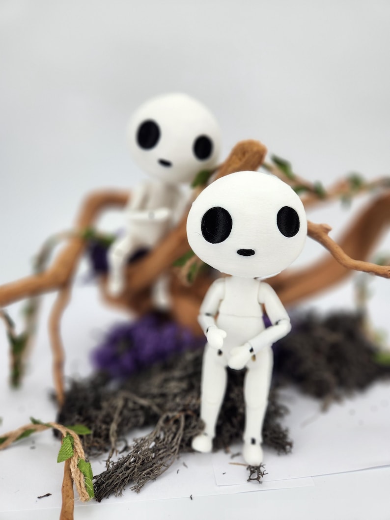 May include: Two white, articulated dolls with black eyes and small mouths, resembling characters from a fantasy film. They are posed in front of a backdrop of twigs, moss, and purple flowers. The dolls have a minimalist design and are set against a blurred background.