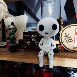 May include: A white articulated figure with large black eyes and a small mouth sits on a wooden shelf. The figure is surrounded by other figurines, books, and decorative items. A circular patch with a red griffin design is visible.