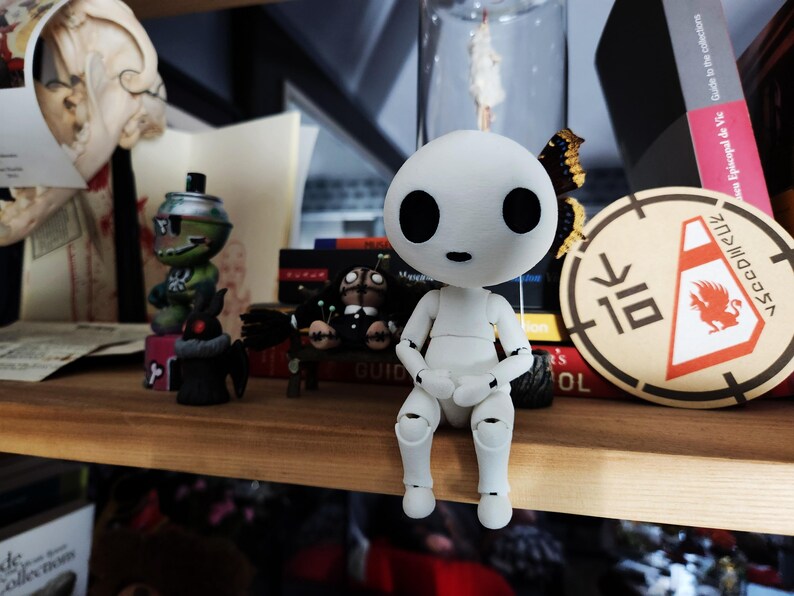 May include: A white articulated figure with large black eyes and a small mouth sits on a wooden shelf. The figure is surrounded by other figurines, books, and decorative items. A circular patch with a red griffin design is visible.