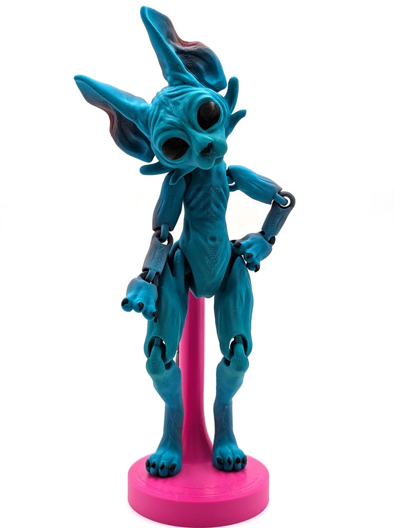 May include: A blue and black articulated doll with large eyes and pointed ears, standing on a pink base.