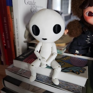 May include: A white articulated doll with large black eyes and a small mouth sits atop a stack of books. The doll has a round head and a simple, minimalist design. The doll is posed with its legs bent and arms crossed.