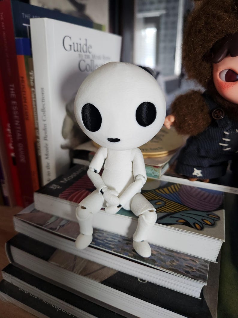 May include: A white articulated doll with large black eyes and a small mouth sits atop a stack of books. The doll has a round head and a simple, minimalist design. The doll is posed with its legs bent and arms crossed.