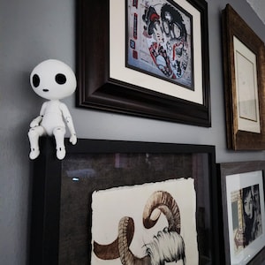 May include: A white, articulated figurine sits atop a framed artwork. The wall displays several framed pieces, including a colorful print with Japanese-inspired imagery and a drawing of a person with large horns. The frames are various sizes and colors.