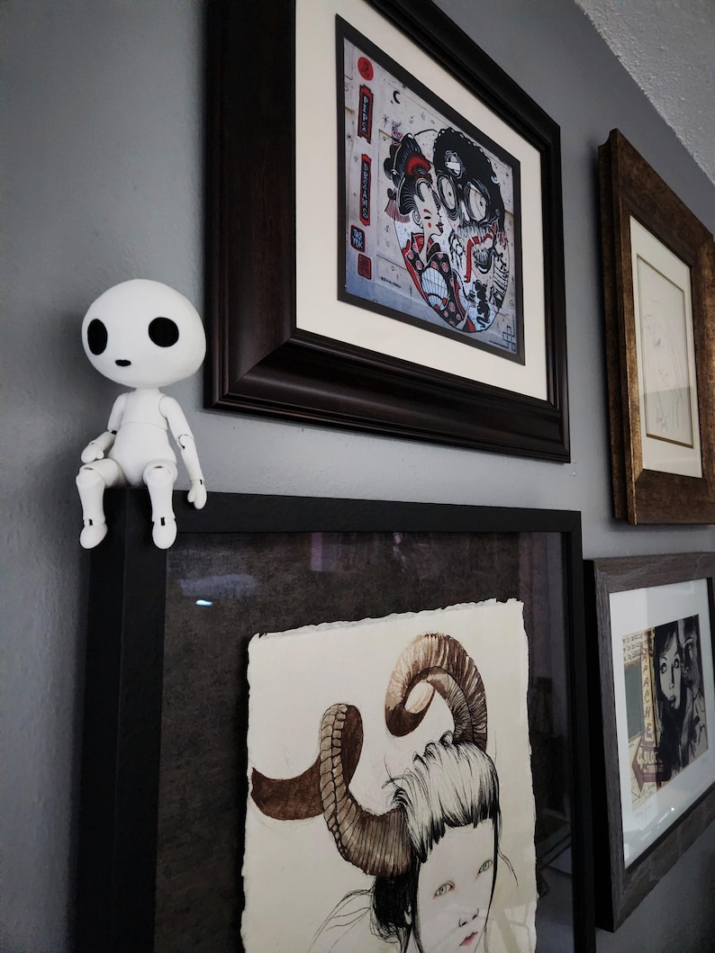 May include: A white, articulated figurine sits atop a framed artwork. The wall displays several framed pieces, including a colorful print with Japanese-inspired imagery and a drawing of a person with large horns. The frames are various sizes and colors.