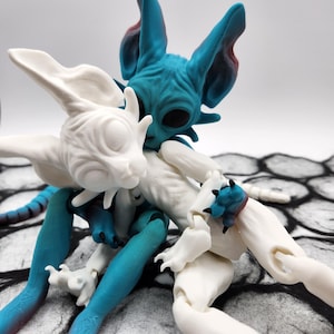 May include: Two articulated cat dolls, one white and one blue, are posed in a hug. The blue cat doll has a textured, wrinkled surface and large, black eyes. The white cat doll has a smooth surface and closed eyes. Both dolls have long, slender limbs and large, pointed ears.