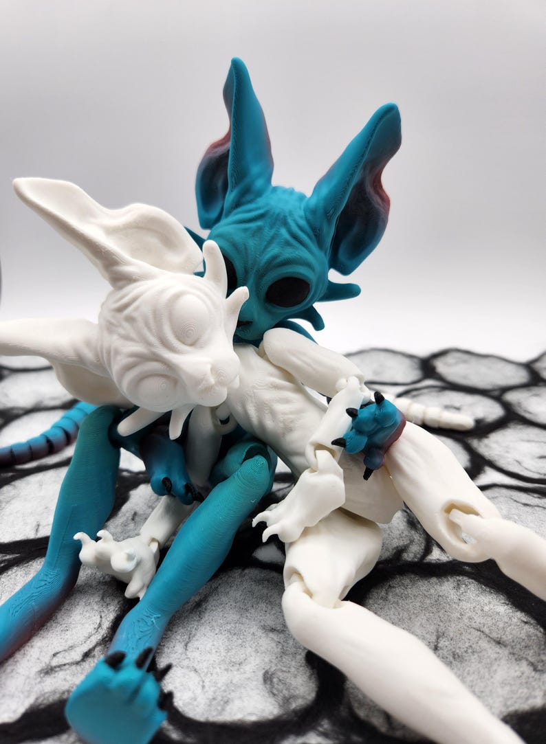 May include: Two articulated cat dolls, one white and one blue, are posed in a hug. The blue cat doll has a textured, wrinkled surface and large, black eyes. The white cat doll has a smooth surface and closed eyes. Both dolls have long, slender limbs and large, pointed ears.
