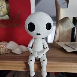 May include: A white articulated doll with a round head and large black eyes sits on a wooden shelf. The doll's arms and legs are poseable, and it is dressed in a simple white outfit. The doll is a unique decorative item.