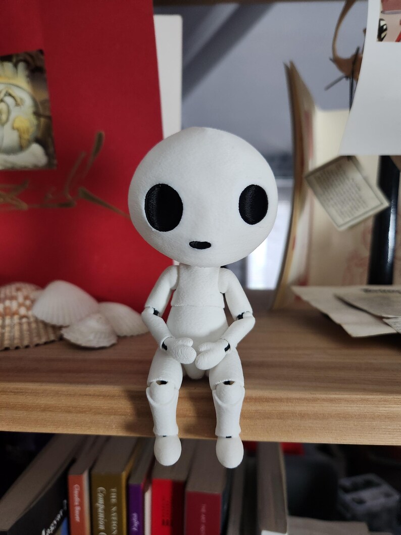May include: A white articulated doll with a round head and large black eyes sits on a wooden shelf. The doll's arms and legs are poseable, and it is dressed in a simple white outfit. The doll is a unique decorative item.